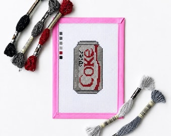May include: A cross-stitch project on white Aida fabric with a pink border, featuring a Diet Coke can design. The embroidery displays the red and silver logo. Five skeins of embroidery floss in black, silver, and red are arranged around the fabric.