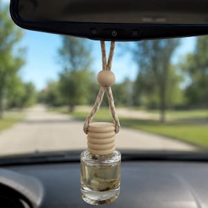 Pode incluir: Um pequeno frasco de vidro transparente preenchido com um líquido claro, com uma tampa de madeira estriada. O frasco pende de um cordão de sisal bege com uma conta de madeira redonda, suspenso no espelho retrovisor de um veículo.
