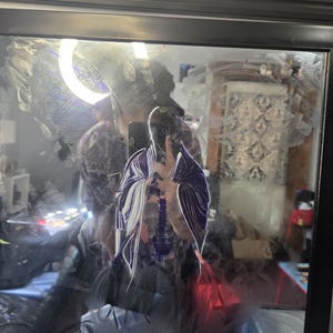 May include: A square wall mirror with a black frame featuring a reflective surface marked by white and purple paint streaks. The glass surface displays smudges and smears of paint, creating a layered visual effect across the reflective plane.