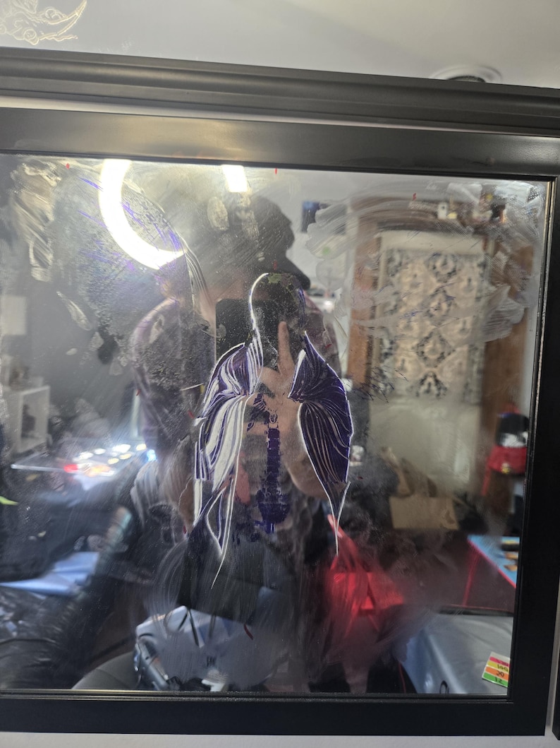 May include: A square wall mirror with a black frame featuring a reflective surface marked by white and purple paint streaks. The glass surface displays smudges and smears of paint, creating a layered visual effect across the reflective plane.