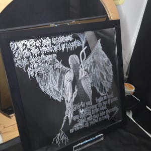 May include: A square black-framed glass art piece featuring white etched line-art of a winged figure with long, flowing hair. The artwork includes intricate white calligraphic text surrounding the central figure, set against a dark background.