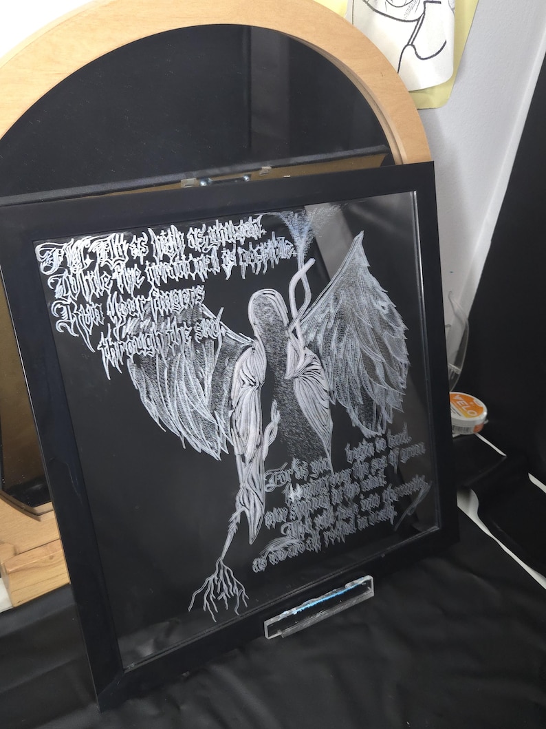 May include: A square black-framed glass art piece featuring white etched line-art of a winged figure with long, flowing hair. The artwork includes intricate white calligraphic text surrounding the central figure, set against a dark background.