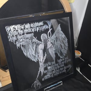 May include: A square black-framed glass art piece featuring white line-art of a winged figure with long hair and intricate text written in a stylized gothic script. The artwork depicts a central figure with large wings and root-like appendages.