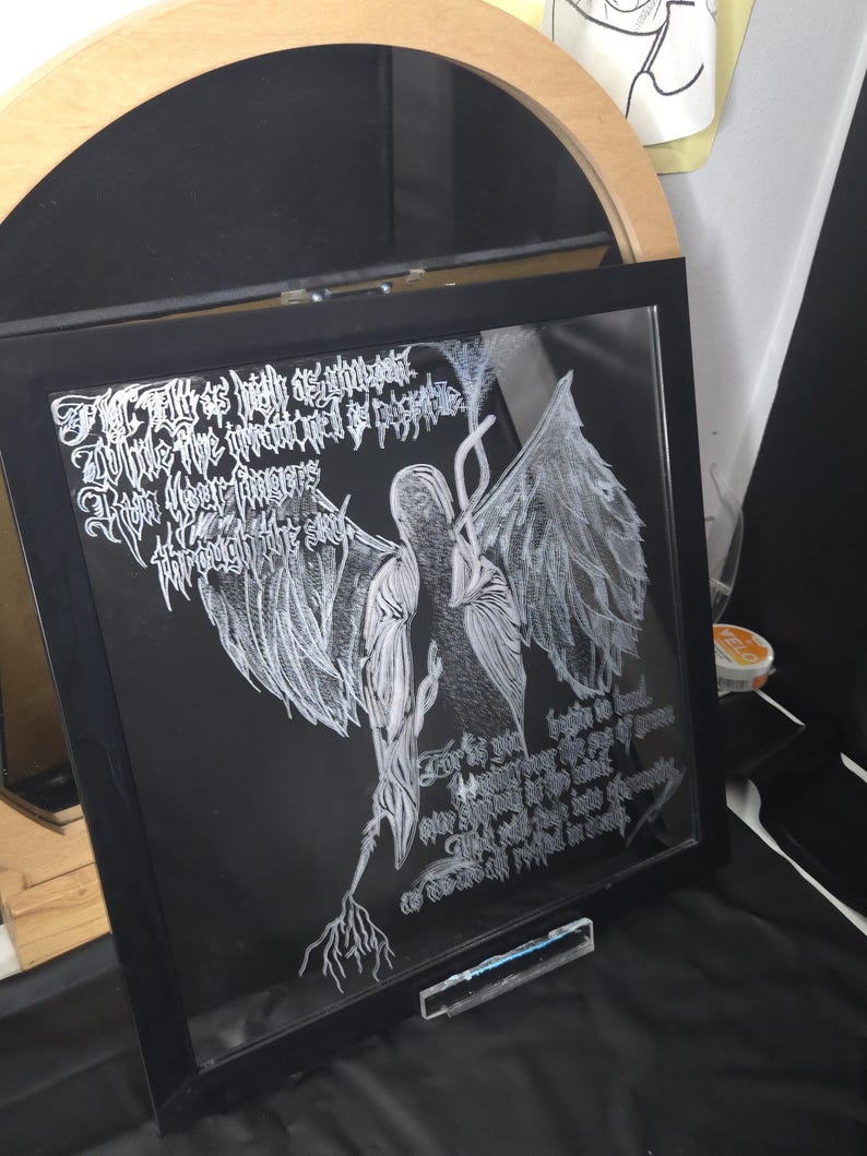 May include: A square black-framed glass art piece featuring white line-art of a winged figure with long hair and intricate text written in a stylized gothic script. The artwork depicts a central figure with large wings and root-like appendages.