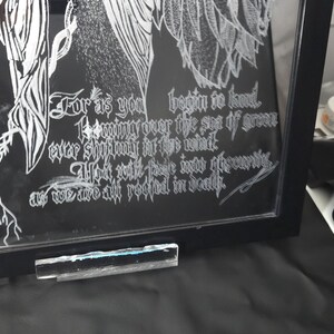 May include: A black framed glass art piece featuring white etched line art of wings and roots. The text reads: For as you begin to kneel, looming over the sea of green ever shifting in the wind, you will fade into obscurity, as we are all rooted in death.