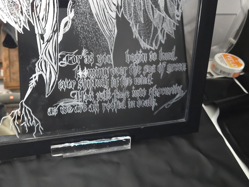 May include: A black framed glass art piece featuring white etched line art of wings and roots. The text reads: For as you begin to kneel, looming over the sea of green ever shifting in the wind, you will fade into obscurity, as we are all rooted in death.
