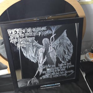 May include: A square black-framed wall art piece featuring a white ink illustration of a winged figure on a black background. The artwork includes intricate white text written in a stylized gothic font, depicting a central figure with large wings.