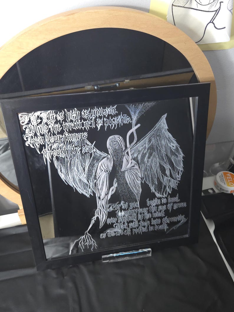 May include: A square black-framed wall art piece featuring a white ink illustration of a winged figure on a black background. The artwork includes intricate white text written in a stylized gothic font, depicting a central figure with large wings.