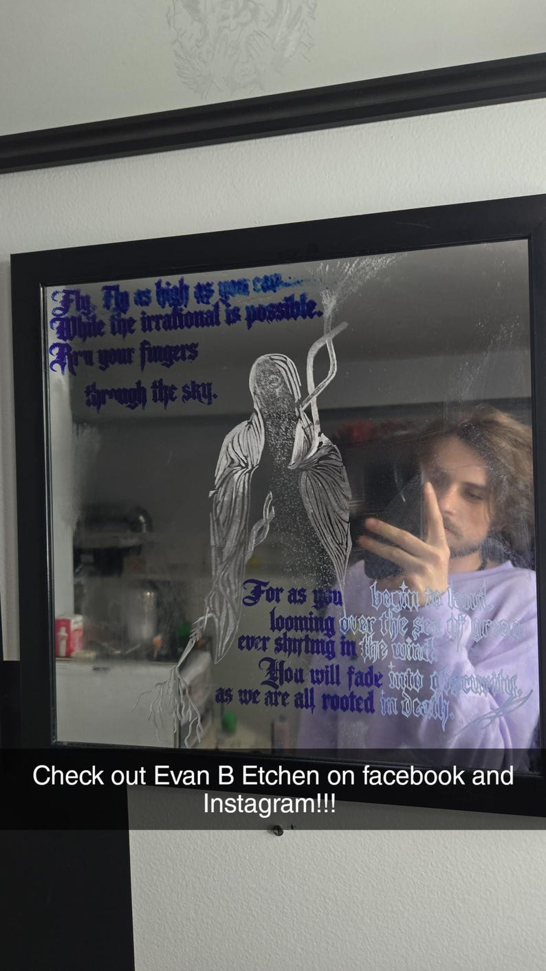 May include: A square wall mirror with a black frame featuring a white line-art illustration of a hooded figure and purple gothic-style text that reads: Fly, Fly as high as you can, While the irrational is possible, Run your fingers through the sky.