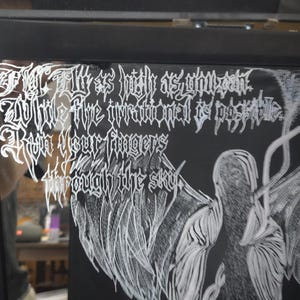 May include: A black framed mirror featuring white etched text that reads: Fly as high as you can, While the irrational is possible, Run your fingers through the sky. Next to the text is a white etched illustration of a winged figure with feathers.