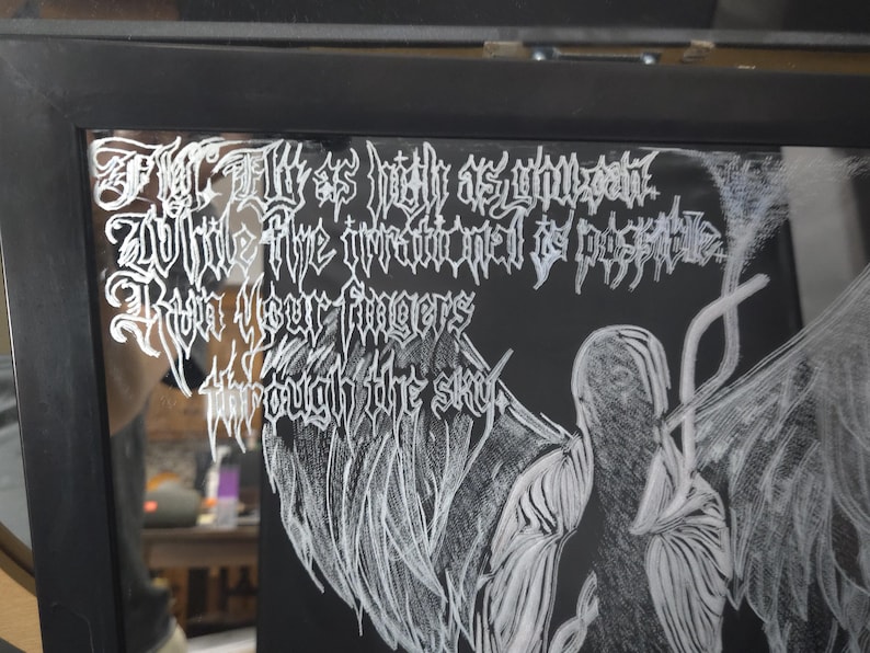 May include: A black framed mirror featuring white etched text that reads: Fly as high as you can, While the irrational is possible, Run your fingers through the sky. Next to the text is a white etched illustration of a winged figure with feathers.