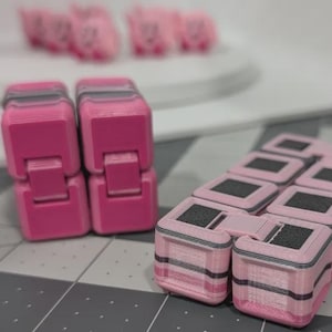 May include: Two solid pink fidget cubes standing upright next to a larger, articulated fidget toy composed of multiple pink and black square segments. The articulated toy features a grid-like pattern of pink and black blocks connected by hinges.