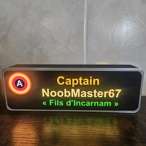 Peut inclure: Un panneau lumineux rectangulaire avec une face noire et un cadre argenté. Le côté gauche présente un logo circulaire avec un A bleu sur fond rouge et jaune. Le texte indique Captain NoobMaster67 et Fils d'Incarnam en vert.