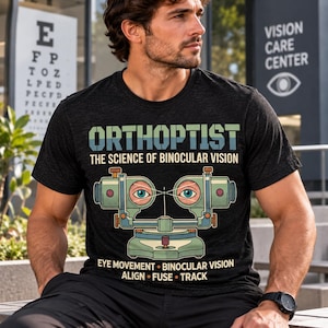 Könnte beinhalten: Ein anthrazitfarbenes T-Shirt mit einer Grafik eines orthoptischen Instruments mit zwei Augen. Der Text lautet: ORTHOPTIST THE SCIENCE OF BINOCULAR VISION, und unter dem Instrument: EYE MOVEMENT BINOCULAR VISION ALIGN FUSE TRACK.
