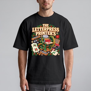 May include: A black t-shirt featuring a graphic illustration of a green letterpress machine surrounded by holiday greenery, red berries, and printing supplies. The text "THE LETTERPRESS PRINTER'S" is printed in large, bold, light-colored letters above the machine.