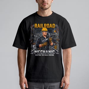 Puede incluir: Una camiseta negra con una ilustración gráfica de un mecánico ferroviario con casco amarillo y guantes, sosteniendo una herramienta eléctrica. El texto dice RAILROAD MECHANIC KEEPING THE RAILS MOVING, con piezas de tren en el fondo.