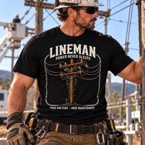 May include: A black t-shirt featuring a graphic of a wooden utility pole with power lines. The text reads LINEMAN POWER NEVER SLEEPS at the top and HIGH VOLTAGE GRID MAINTENANCE at the bottom in white and orange lettering on the front of the shirt.