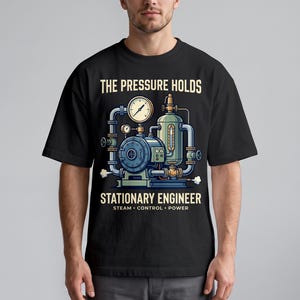 May include: A black short-sleeve t-shirt featuring a graphic of industrial machinery with pipes and a pressure gauge. The text reads THE PRESSURE HOLDS STATIONARY ENGINEER STEAM CONTROL POWER in a light yellow font centered on the chest.