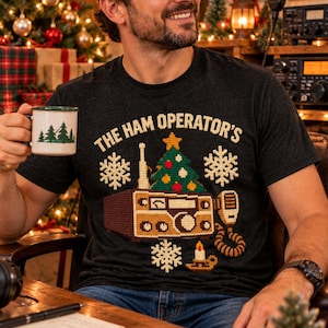 Op de afbeelding: Een donkergrijs T-shirt met een grafische print van een vintage radio met een kerstboom erop, omringd door sneeuwvlokken en een kaars. De tekst "THE HAM OPERATOR'S" is gedrukt in een lichtgeel, gestructureerd lettertype in kruissteekstijl.