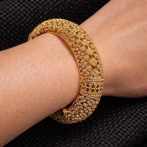 Puede incluir: Un brazalete rígido de oro con un intrincado diseño de celosía perforada y pequeñas texturas en relieve similares a cuentas en toda la superficie. La banda circular ancha tiene un acabado pulido y una construcción con bisagras para el cierre.
