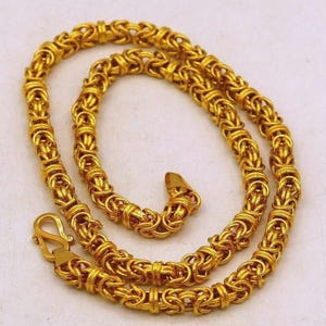 Puede incluir: Un collar de cadena de color dorado con un patrón complejo de eslabones bizantinos entrelazados. El collar está construido con una serie de bucles de metal tejidos y repetitivos, e incluye un cierre de gancho en forma de S en un extremo.