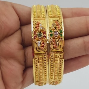 Puede incluir: Dos brazaletes de oro con intrincados patrones de filigrana, paneles rectangulares texturizados y motivos florales acentuados con pequeñas piedras verdes, rojas y transparentes. La superficie metálica muestra un acabado pulido.