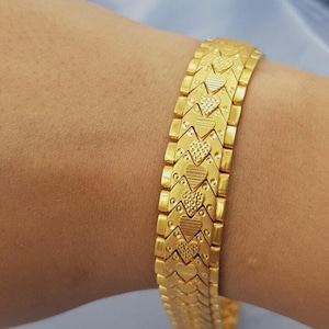 Puede incluir: Una pulsera de tono dorado que presenta un patrón repetitivo de eslabones entrelazados en forma de corazón. Cada eslabón de corazón muestra una superficie texturizada con líneas horizontales alternas y detalles punteados, conectados por segmentos laterales rectangulares lisos.