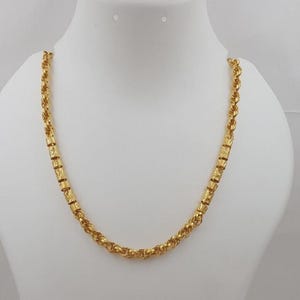 Puede incluir: Un collar de cadena dorada que presenta un patrón repetitivo de eslabones cilíndricos texturizados intercalados con complejos eslabones de estilo cuerda trenzada. El acabado amarillo metálico tiene un brillo pulido, creando un diseño continuo y uniforme en toda la longitud de la pieza de joyería.