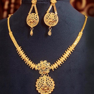 Puede incluir: Un conjunto de joyería en oro que consta de un collar y dos pendientes a juego. El collar presenta un motivo floral central con un colgante circular de filigrana y una cadena de cuentas texturizadas. Los pendientes reflejan el diseño de filigrana.