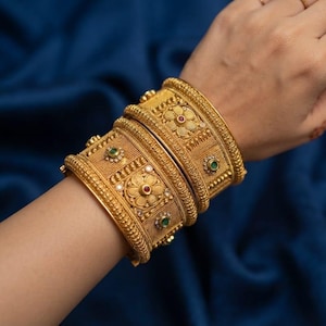 Pode incluir: Dois braceletes largos de ouro com intrincados padrões florais em relevo, texturas granuladas e pequenas pedras preciosas vermelhas e verdes incrustadas. Os braceletes têm um design espesso e estruturado com bordas de metal detalhadas.