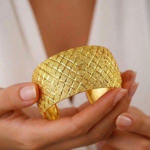 Puede incluir: Una pulsera brazalete ancha de oro con un patrón de rejilla texturizado en forma de diamante en toda su superficie exterior. El metal tiene un acabado mate y granulado que capta la luz, y el brazalete presenta un diseño abierto en la parte posterior.