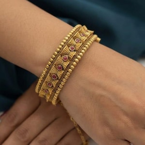 Puede incluir: Un brazalete dorado con una banda central de motivos repetitivos en forma de abanico, cada uno con una pequeña piedra roja redonda. El brazalete está bordeado por dos filas de detalles metálicos dorados texturizados y con cuentas en los bordes.