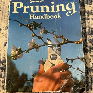 May include: A softcover book titled Sunset Pruning Handbook with a blue cover featuring a photograph of a hand using metal pruning shears to trim a flowering branch against a clear blue sky. The cover shows signs of wear and creasing.