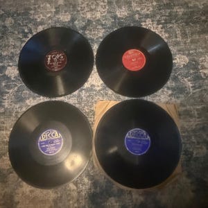 May include: Four black vinyl phonograph records arranged in a square formation. The top left record has a maroon Tops label, the top right has a red Columbia label, and the bottom two have blue Decca labels with visible text and song titles.