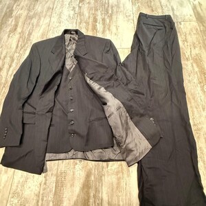 May include: A dark charcoal gray three-piece suit consisting of a blazer, a button-front vest, and matching trousers. The fabric features a subtle pinstripe pattern, and the garments are lined with a smooth, solid gray interior fabric.