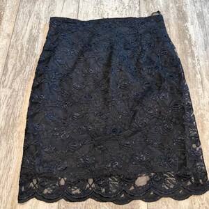 May include: A black knee-length skirt featuring an intricate floral lace overlay pattern. The garment has a high waistline and a scalloped hem edge that follows the floral design. The lace fabric is semi-sheer, revealing a solid black lining underneath.