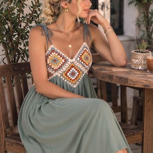 May include: A midi-length dress featuring a sage green skirt and a bodice constructed from multi-colored crochet granny squares in shades of orange, yellow, white, and grey. The garment includes thin, adjustable tie-shoulder straps and a V-neckline.