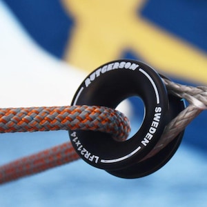Peut inclure: Un anneau à faible friction noir et circulaire Rutgerson Sweden LFR21x14 en métal, avec du texte blanc imprimé sur sa surface. Une corde tressée grise et orange passe par le centre de l'anneau, tandis qu'une corde grise est passée en boucle.