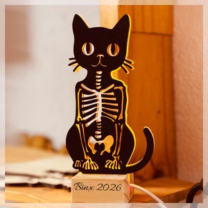 May include: A black cat-shaped wooden lamp with a cutout skeleton design, illuminated from behind with a warm glow. The base is a light-colored wooden block featuring the engraved text "Binx 2026" in a cursive font, with a white power cord attached.