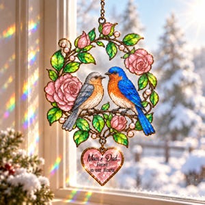 May include: A stained glass suncatcher featuring two bluebirds perched on a branch within a circular wreath of pink roses and green leaves. A heart-shaped pendant hangs from the bottom with the text Mom & Dad Forever in our Hearts.