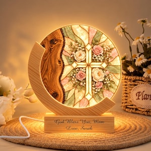 May include: A circular illuminated lamp featuring a stained glass design with a central cross surrounded by pink and white roses. The lamp is set in a crescent-shaped wooden frame on a rectangular base engraved with "God Bless You, Mam Love, Sarah."