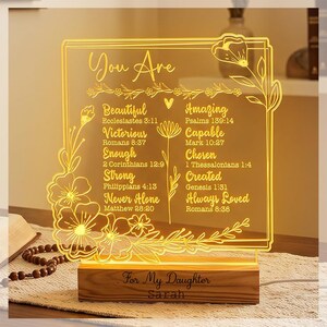 May include: A square acrylic light plaque with warm yellow illumination featuring floral engravings and inspirational text including "You Are Beautiful," "Amazing," "Victorious," "Capable," "Enough," "Chosen," "Strong," "Created," "Never Alone," and "Always Loved" with Bible verses, set on a wooden base engraved with "For My Daughter" and "Sarah."