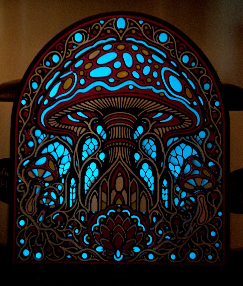 May include: An arched wall art piece featuring a layered design of mushrooms and intricate patterns. The artwork displays a dark frame with cutouts that reveal a vibrant, glowing cyan light behind the red and black detailed mushroom motifs.
