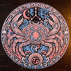 Może przedstawiać: Okrągła ozdobna tabliczka z centralną ilustracją kraba otoczoną misternymi zawijasami, p&oacute;łksiężycem i symbolem zodiakalnego Raka. Wz&oacute;r wykorzystuje paletę kolor&oacute;w składającą się z czerni, jasnego błękitu i delikatnego r&oacute;żu.