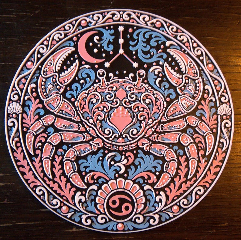 Może przedstawiać: Okrągła ozdobna tabliczka z centralną ilustracją kraba otoczoną misternymi zawijasami, p&oacute;łksiężycem i symbolem zodiakalnego Raka. Wz&oacute;r wykorzystuje paletę kolor&oacute;w składającą się z czerni, jasnego błękitu i delikatnego r&oacute;żu.