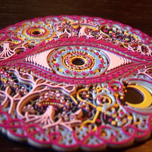 May include: A circular multi-layered art piece featuring a central eye motif surrounded by intricate patterns of trees, a crescent moon, and geometric shapes in vibrant shades of magenta, light blue, yellow, and white with black accents.