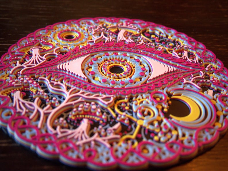 May include: A circular multi-layered art piece featuring a central eye motif surrounded by intricate patterns of trees, a crescent moon, and geometric shapes in vibrant shades of magenta, light blue, yellow, and white with black accents.
