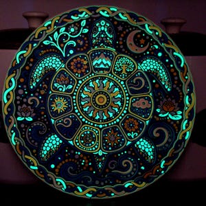 May include: A circular decorative plate featuring a detailed illustration of a sea turtle. The shell is divided into segments containing symbols like a sun, moon, mountains, and flowers. The design glows in the dark with bright cyan accents.