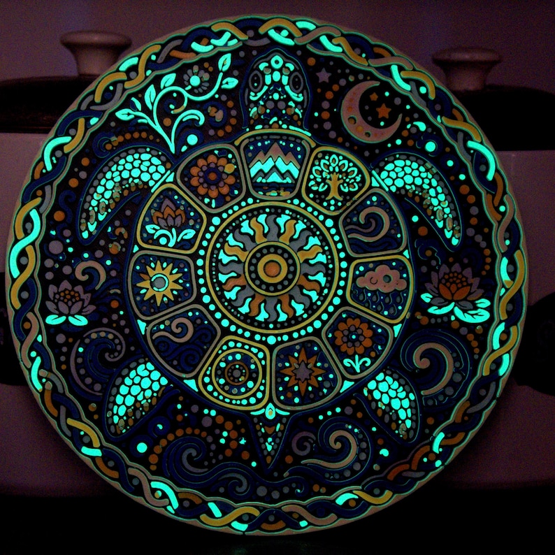 May include: A circular decorative plate featuring a detailed illustration of a sea turtle. The shell is divided into segments containing symbols like a sun, moon, mountains, and flowers. The design glows in the dark with bright cyan accents.