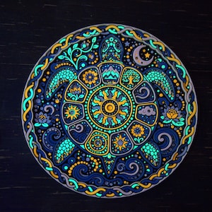 May include: A circular wall art piece featuring a central sea turtle with a shell divided into segments containing sun, mountain, tree, and floral motifs. The design uses a vibrant palette of glowing blue, yellow, and purple against a dark background.